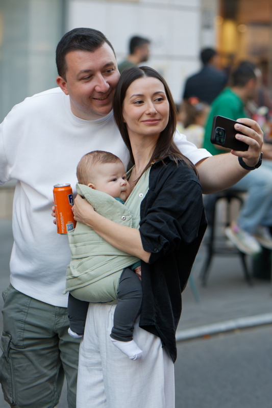 3 - A man in a white sweatshirt and a woman wearing a black shirt over a green baby wrap, holding a baby and taking a selfie together outdoors